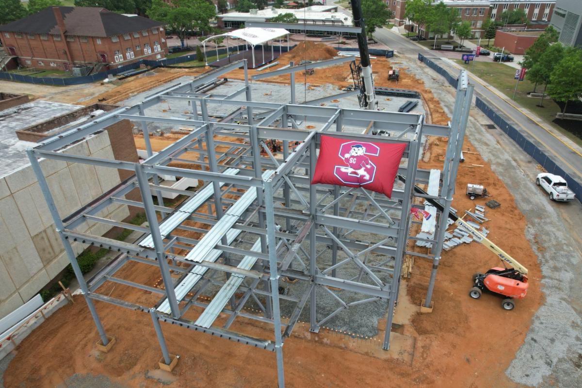 Steel building framework under construction, with a red flag displaying a large "S" and bulldog logo. Construction vehicles and materials are visible on the dirt ground.