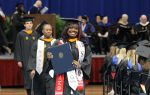 A graduate in a cap and gown smiles, holding a diploma during a graduation ceremony, with other graduates and faculty in the background.