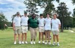 Six people in golf attire posing together on a golf course with trees and a flag in the background.