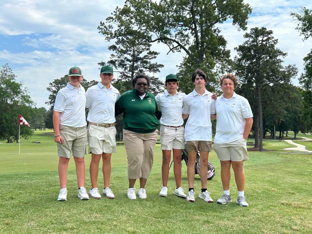 Six people in golf attire posing together on a golf course with trees and a flag in the background.