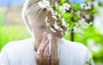 A person outdoors holds a tissue to their nose, appearing to sneeze near blooming white flowers, suggesting seasonal allergies.