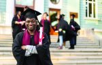 A smiling graduate in a cap and gown holds a diploma, standing in front of a building with other graduates in the background.