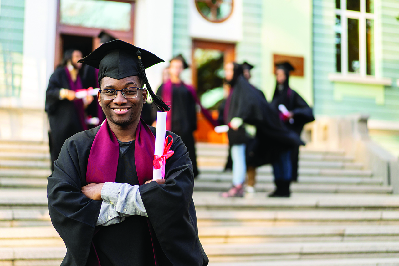 A smiling graduate in a cap and gown holds a diploma, standing in front of a building with other graduates in the background.