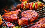 Steaks and colorful vegetable skewers grilling on a barbecue, surrounded by fresh asparagus and a jar of lemonade in the background outdoors.