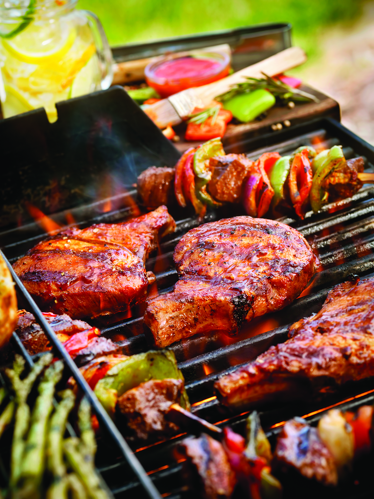 Steaks and colorful vegetable skewers grilling on a barbecue, surrounded by fresh asparagus and a jar of lemonade in the background outdoors.