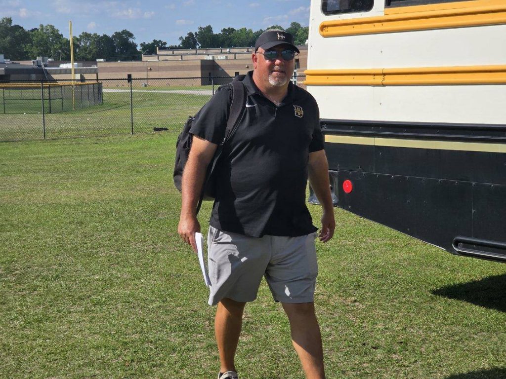 A man wearing sunglasses, a black polo shirt, cap, and shorts, carrying a backpack and papers, walks beside a school bus on a grassy field.