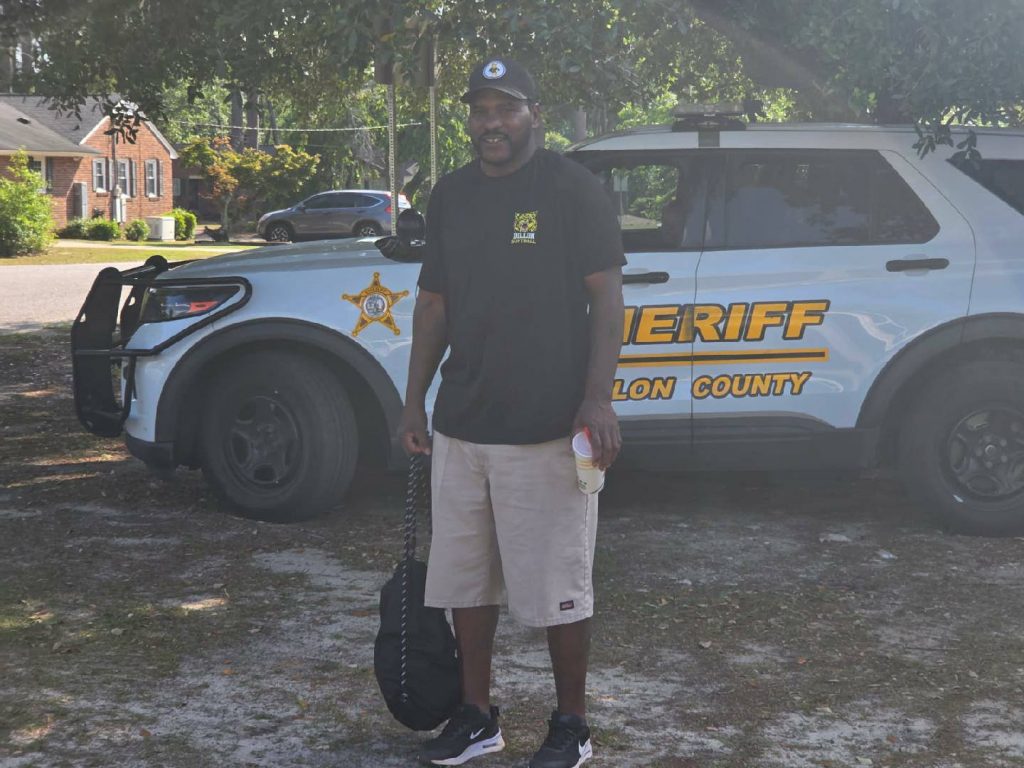 A man stands in front of a sheriff’s vehicle holding a cup and bag, outdoors on a sunny day with trees and houses in the background.