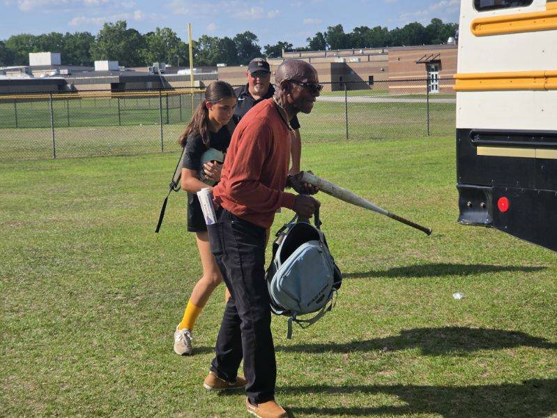 Three people walking toward a yellow school bus on a grassy field, one carrying an umbrella and bag, with a school building in the background.