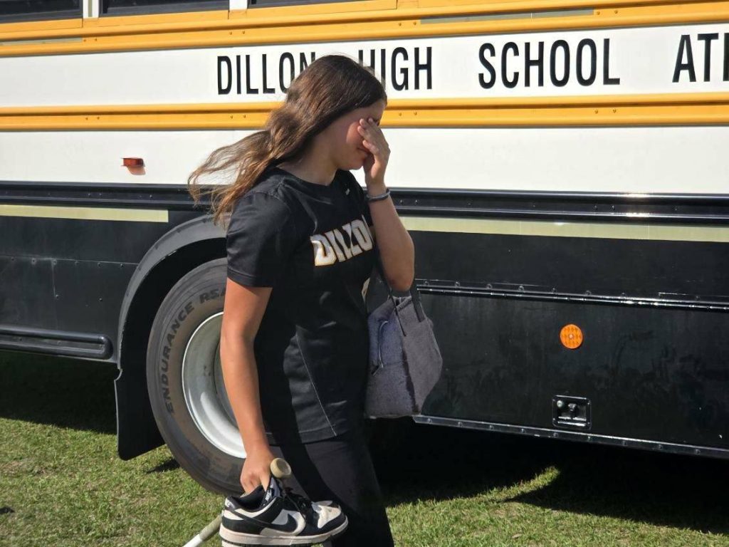 A girl in a black shirt stands in front of a school bus, holding shoes and a bag, shielding her face from the sun.