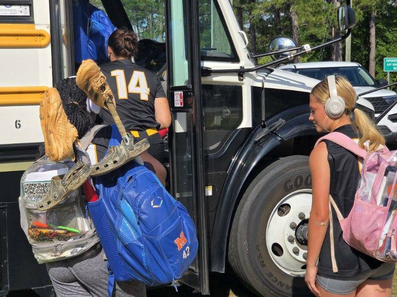 Girls in sports uniforms with backpacks and gear board a school bus; one wears headphones, another carries cleats and a bat.