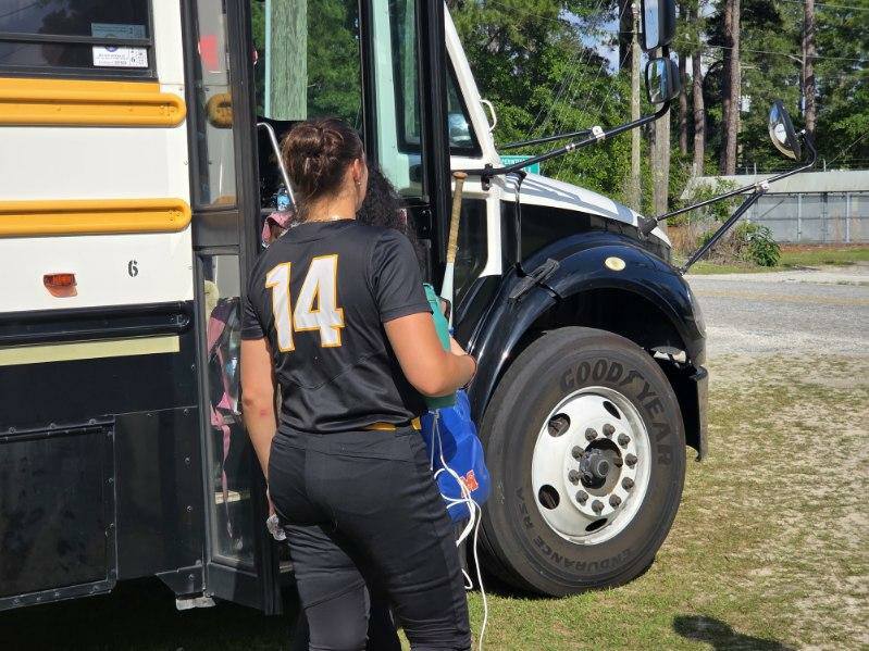 A person in a black uniform with number 14 boards a yellow school bus, holding a blue bag on a sunny day.