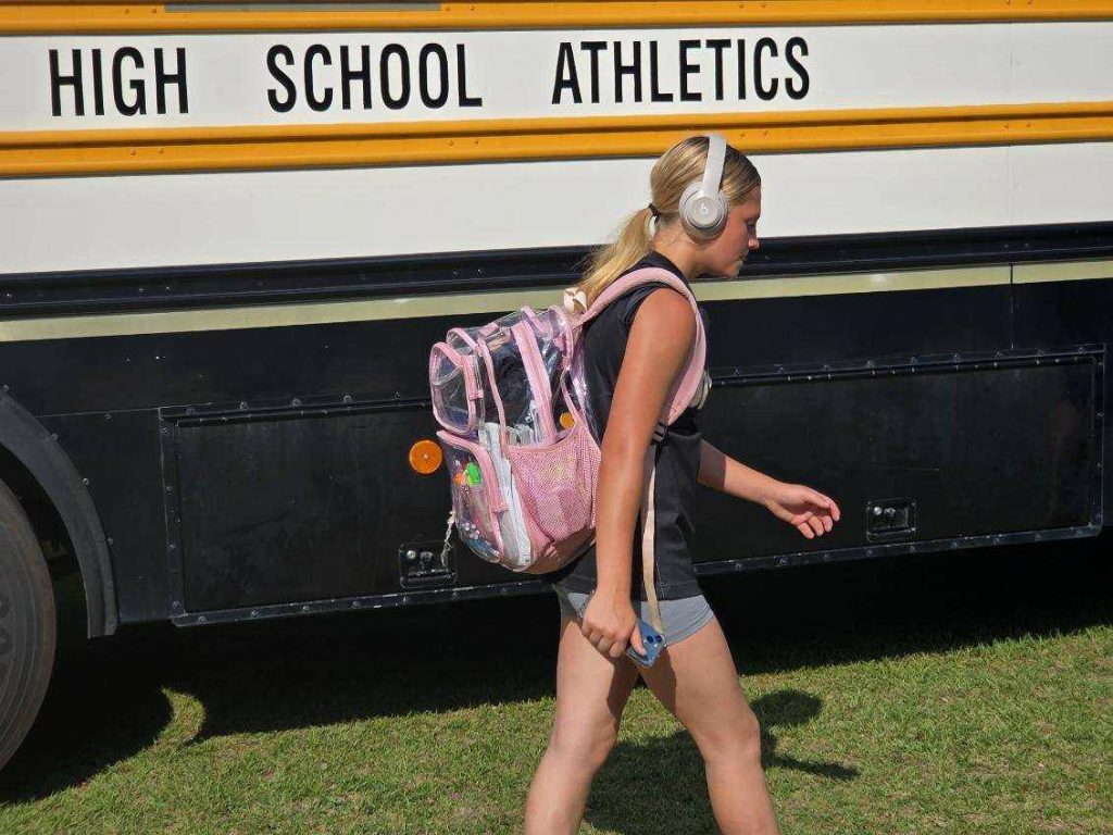 A girl with headphones and a pink backpack walks past a school bus labeled "High School Athletics" on a sunny day.