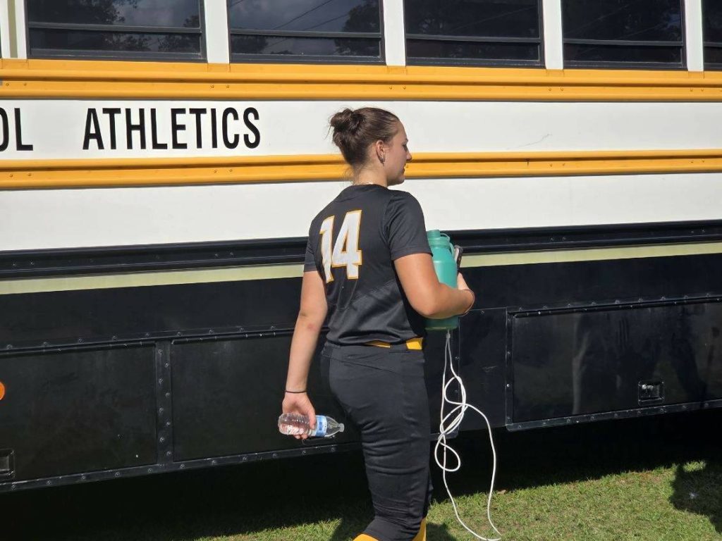 A softball player in uniform number 14 walks beside a school athletics bus, holding a water bottle and a jug, on a sunny day.