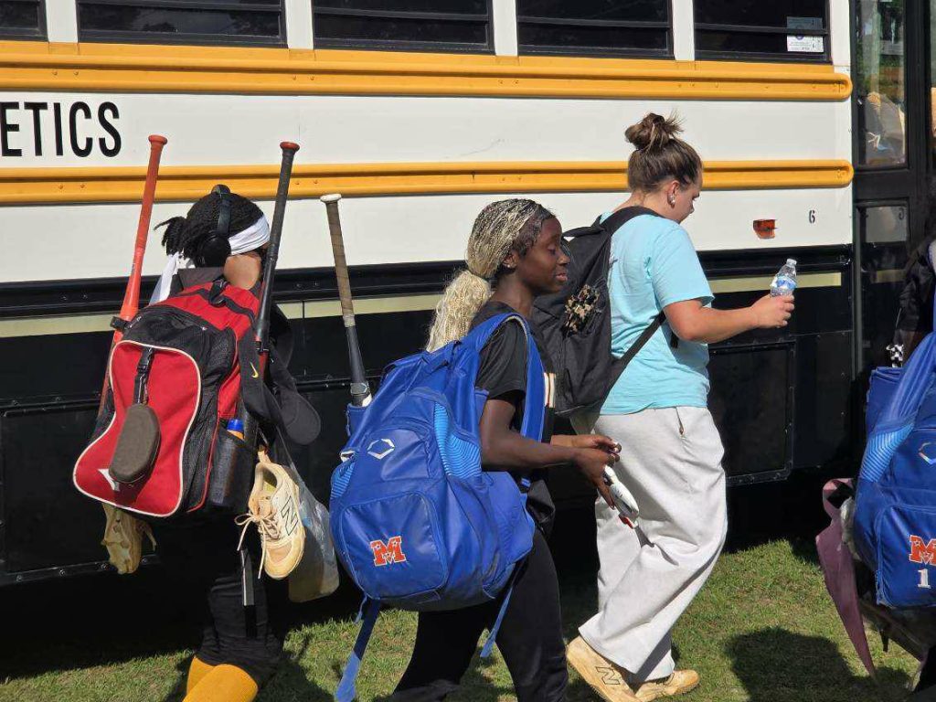 Three girls with sports equipment and backpacks walk toward a yellow school bus on a sunny day.