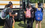 Female athletes with backpacks boarding a yellow school bus; one holds a drink, another carries cleats tied to her blue backpack.