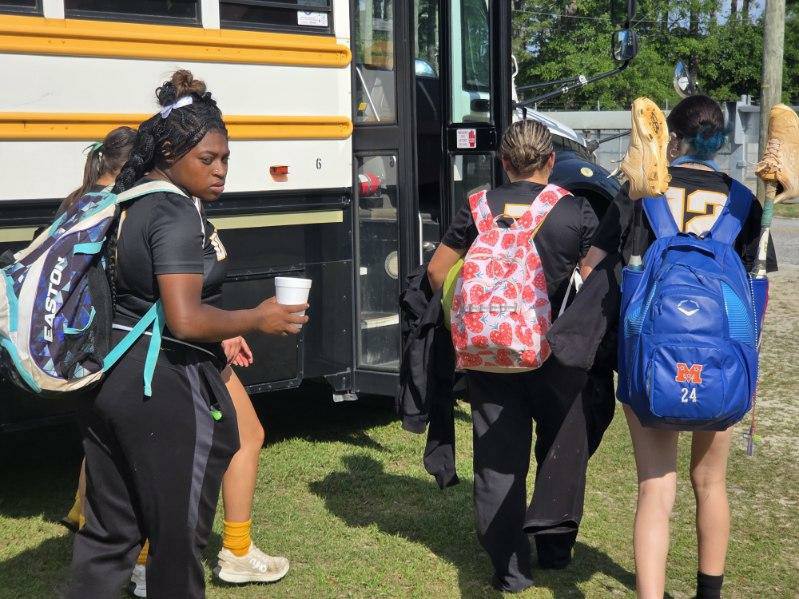 Female athletes with backpacks boarding a yellow school bus; one holds a drink, another carries cleats tied to her blue backpack.