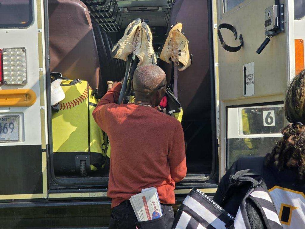 A person loads sports bags and a pair of shoes into the back of a bus. A softball-themed bag and two people are visible.