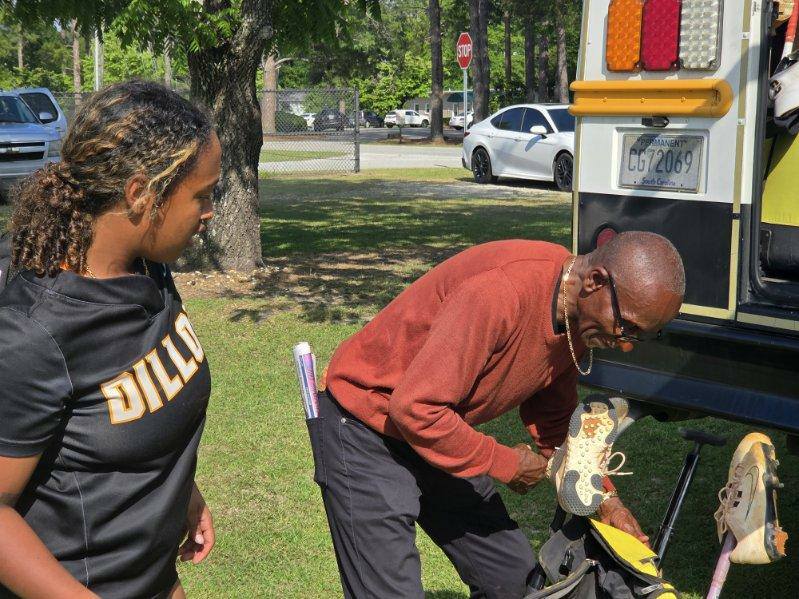 A young athlete in a "DILLON" jersey stands outside as an older man organizes sports gear near an open vehicle trunk.