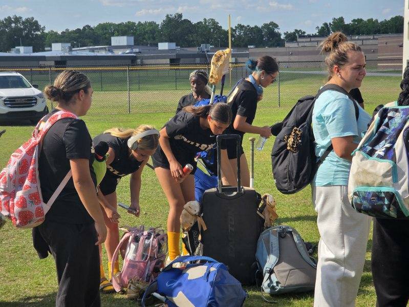 A group of young women with sports bags and equipment stand and prepare on a grassy field, near a fence and school buildings in the background.