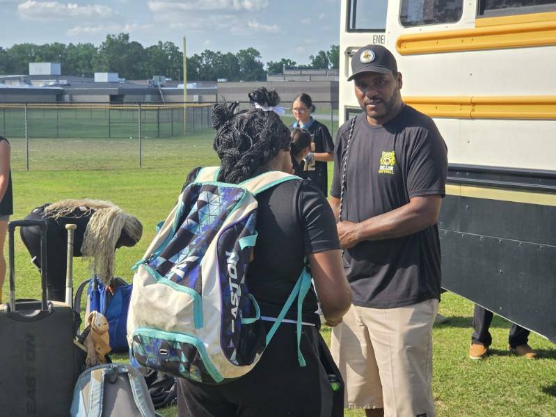 A man and a girl with backpacks stand beside a school bus on a grassy field, surrounded by luggage, on a sunny day.