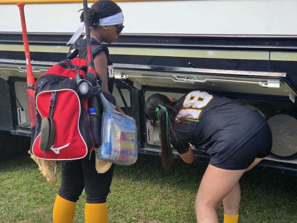 Two female softball players in uniform load equipment into a bus storage compartment. One carries a red bag with bats, while the other arranges items.