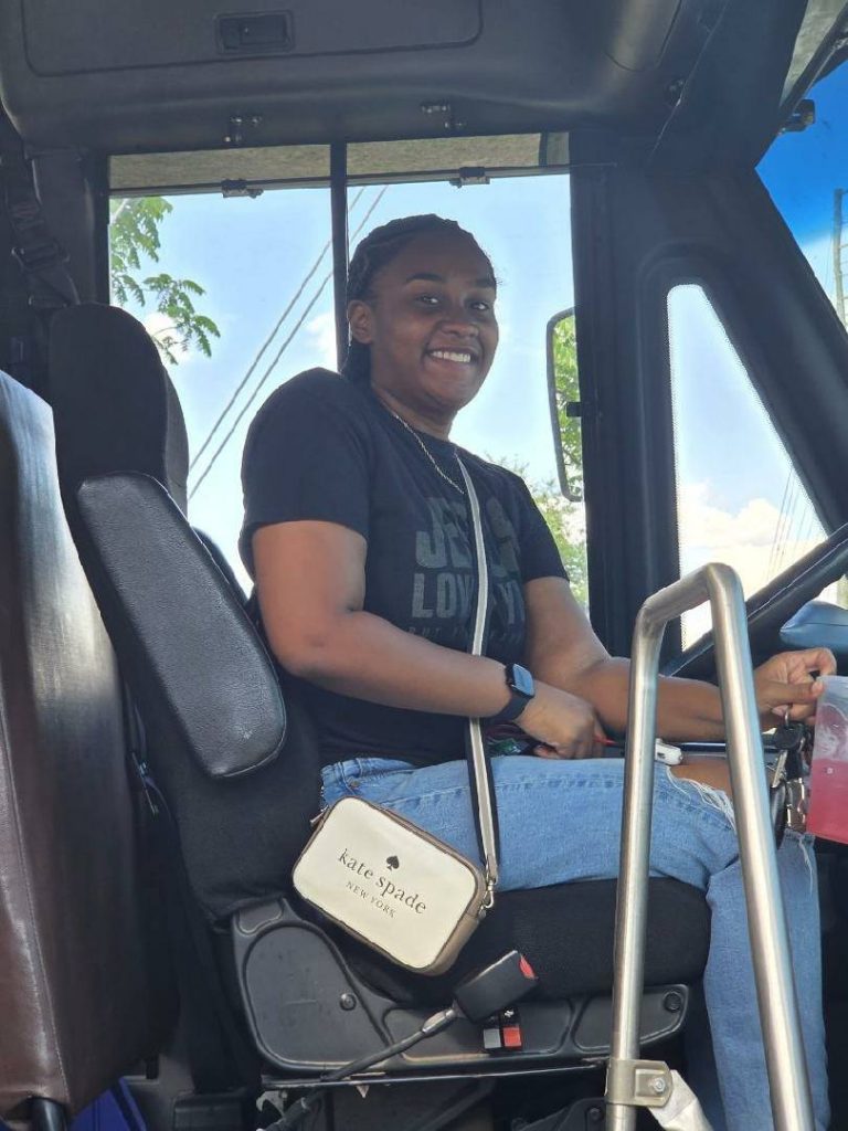 Smiling woman sitting in the driver’s seat of a vehicle, wearing a black t-shirt and jeans, holding a drink and a key, with a Kate Spade purse.