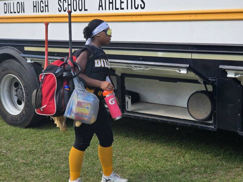 A softball player in uniform and sunglasses carries gear and a drink, walking near a bus labeled "Dillon High School Athletics."