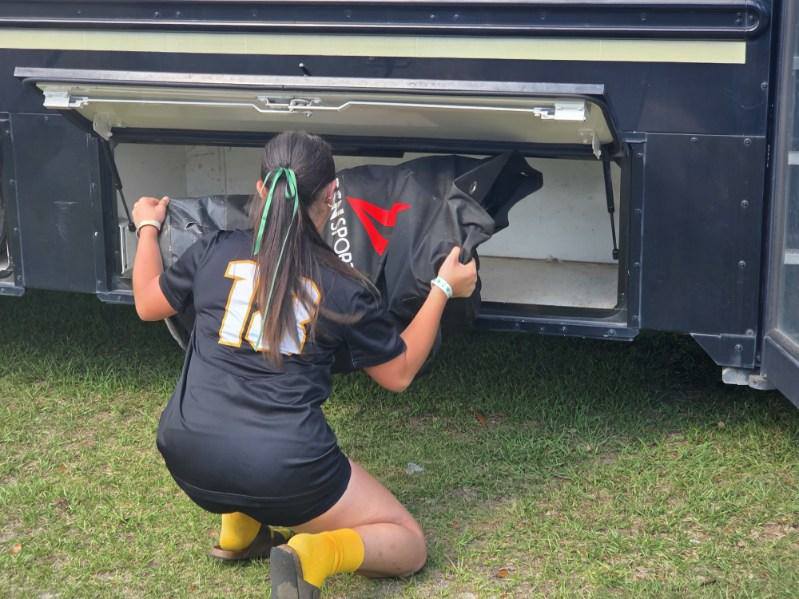 A person in a sports uniform places a large black bag into the storage compartment of a bus, kneeling on grass.