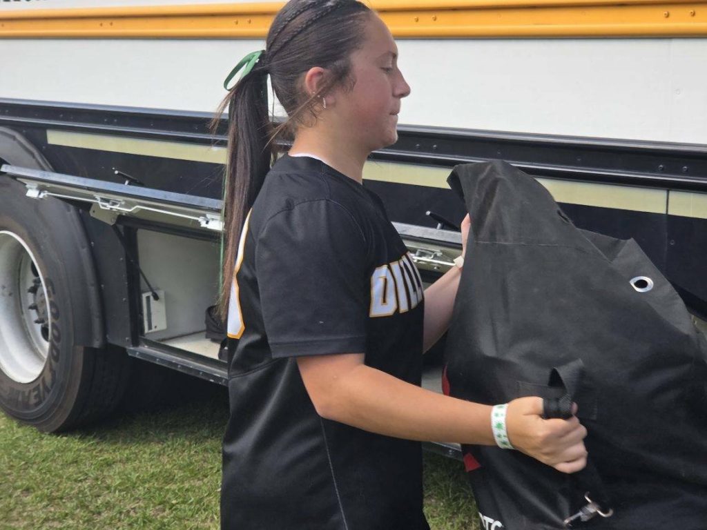 Girl in sports uniform loading a large black bag into a bus storage compartment, standing on grass outdoors.