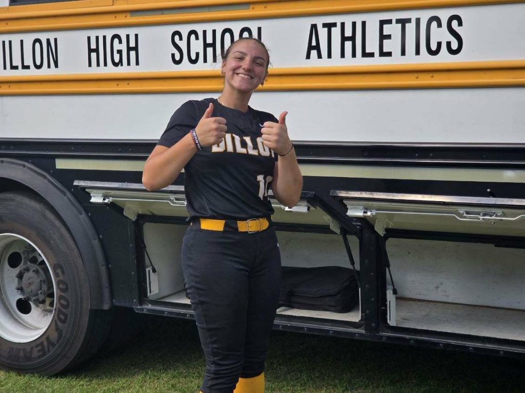 A smiling athlete in a black uniform gives two thumbs up in front of a Dillon High School Athletics bus.