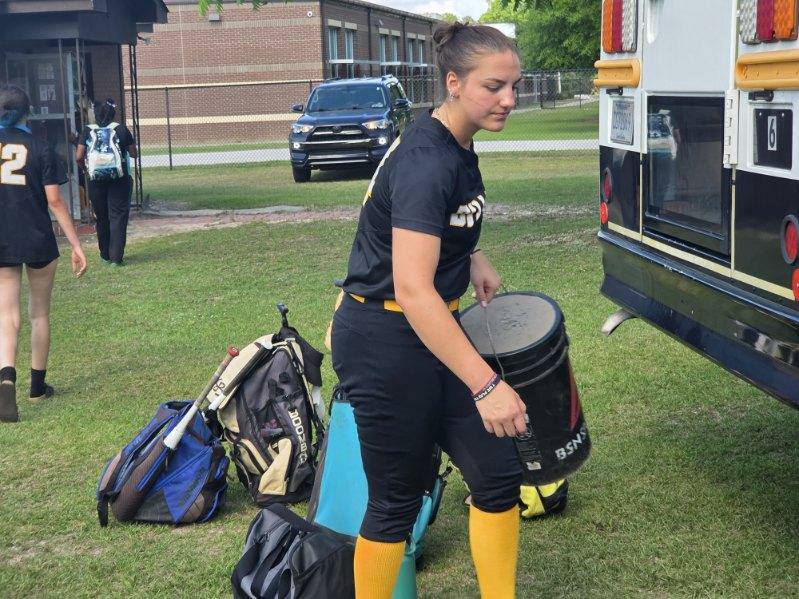 Softball player carrying equipment near a school bus, with other players and sports bags visible on the grass.
