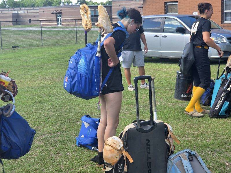Softball players with gear bags and equipment gather on a grassy field near a parked car and building.