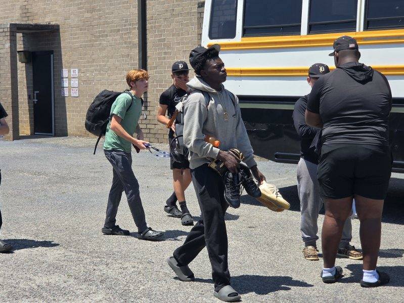 A group of young men in casual sportswear stand and walk near a parked bus on a sunny day, carrying athletic equipment.