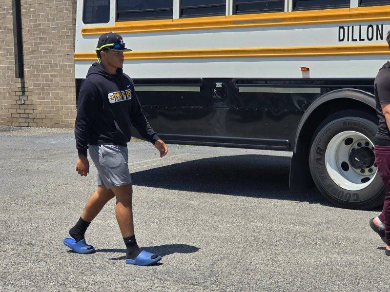 A person in athletic wear and slides walks past a black and yellow school bus parked on a paved lot.