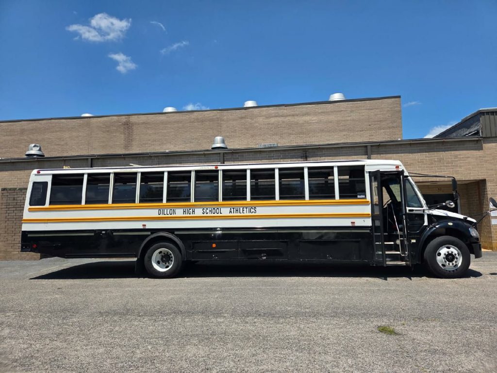 A black and white school bus labeled "Dillon High School Athletics" is parked outside a tan brick building under a clear blue sky.