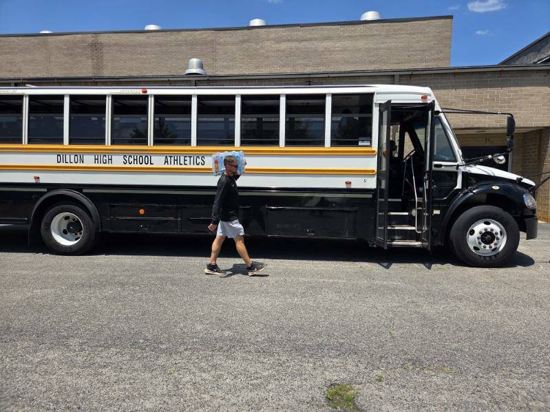 A man walks in front of a Dillon High School Athletics bus parked outside on a sunny day.