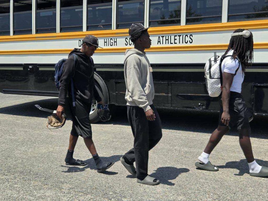 Three young men walk beside a yellow school bus labeled "High School Athletics," carrying backpacks and sports equipment on a sunny day.