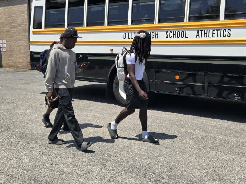 Two young men walk past a school bus labeled "Dillon High School Athletics," one wearing headphones and a backpack, the other looking at his phone.