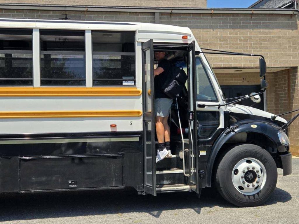 A person wearing shorts and a backpack is standing in the doorway of a school bus parked outside a building.