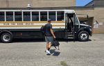 A person carrying bags walks toward a parked school bus labeled "Dillon High School Athletics" on a sunny day.