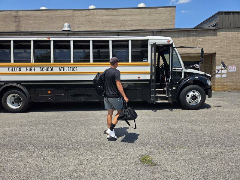 A person carrying bags walks toward a parked school bus labeled "Dillon High School Athletics" on a sunny day.