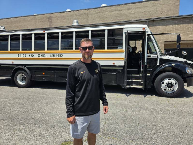 A man in sports attire stands in front of a Dillon High School Athletics bus parked outside a brick building on a sunny day.
