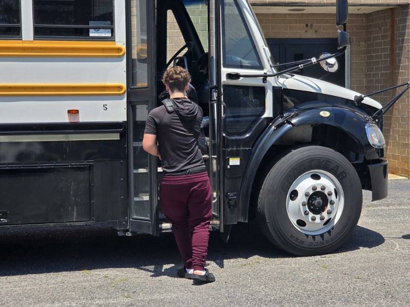 A person wearing a black shirt and red pants steps onto a yellow and black school bus parked outside a brick building.