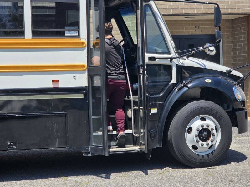 A person wearing maroon pants and a black shirt is boarding a black and white school bus parked near a brick building.
