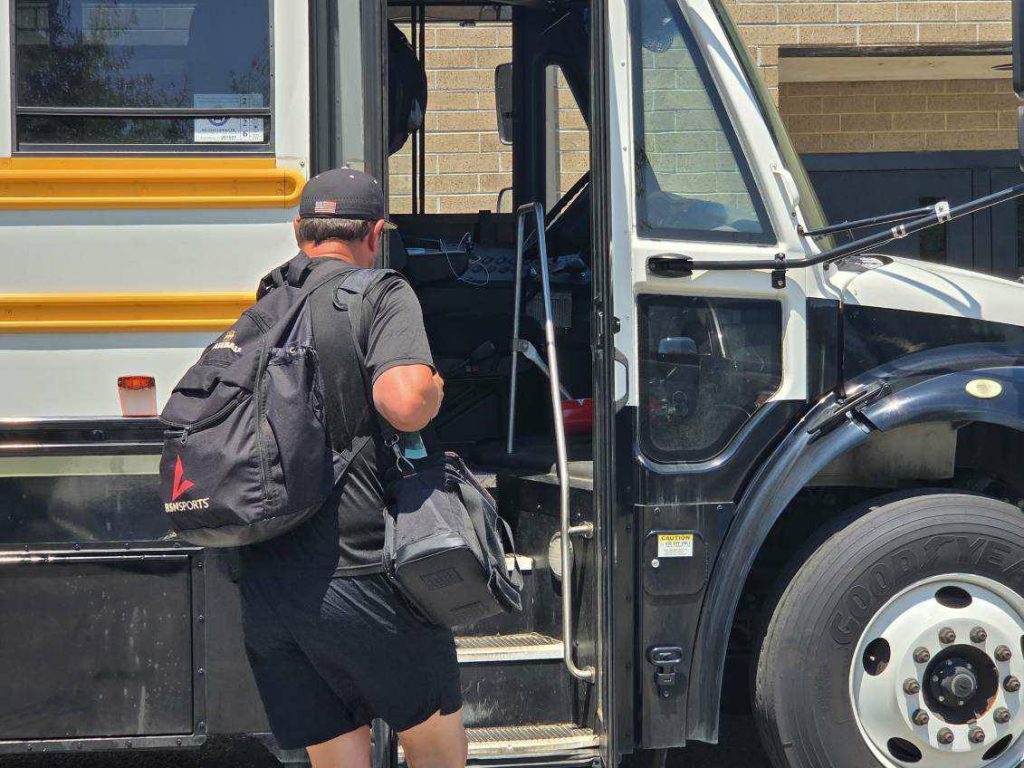 A person with backpacks and a cap is boarding a school bus, holding a duffel bag, with a brick building in the background.