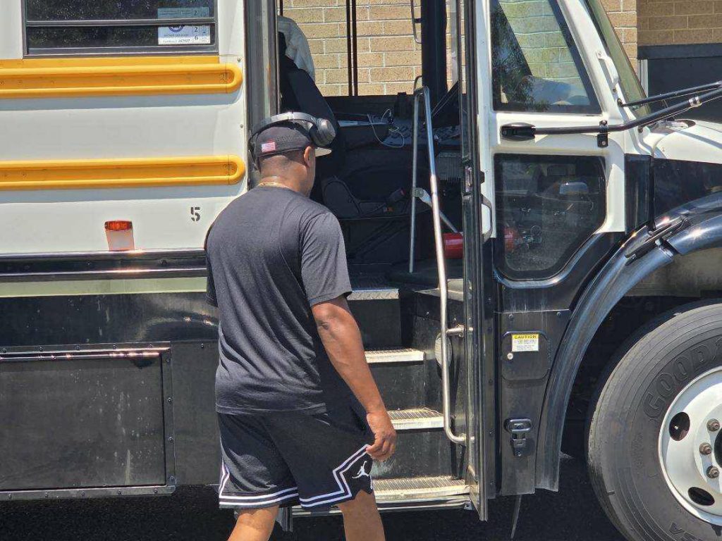 A man wearing headphones and black clothing stands near the open door of a school bus on a sunny day.