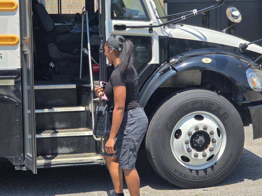 A woman wearing black stands outside a bus, holding keys and a bag, facing the bus steps under bright sunlight.