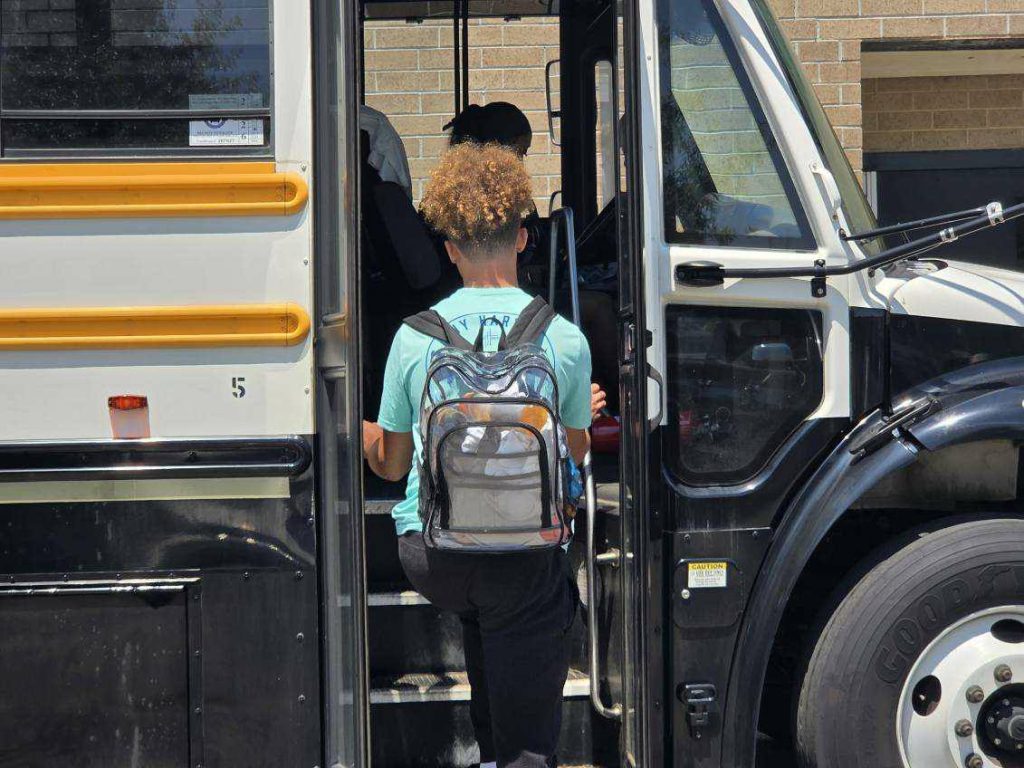 A student with a backpack is boarding a yellow school bus, seen from behind on a sunny day near a brick building.