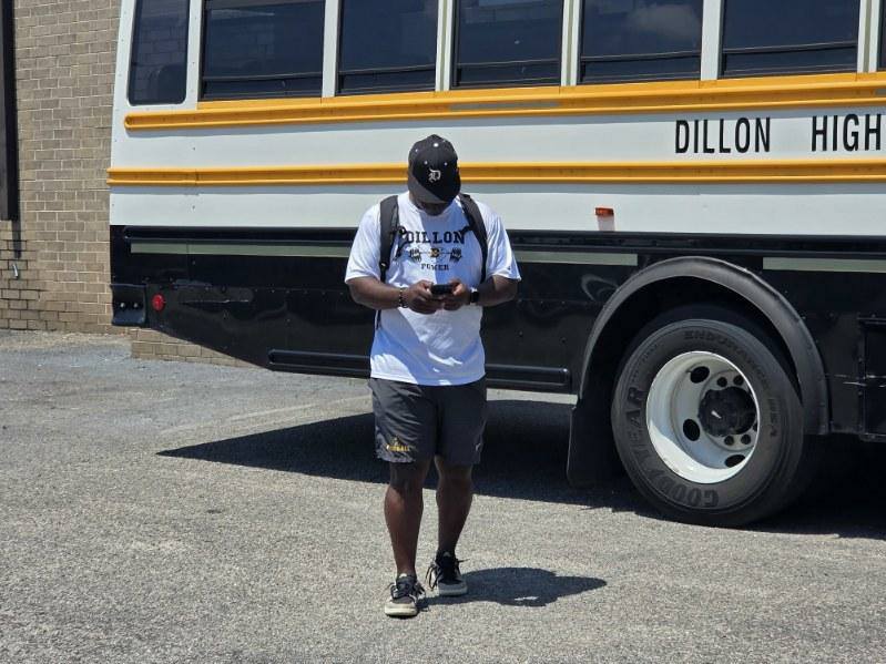 A man wearing a "Dillon High" shirt and hat stands in front of a school bus, looking at his phone.