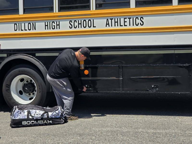 A person in sportswear loads equipment into the storage compartment of a Dillon High School Athletics bus, with a large duffle bag on the ground nearby.